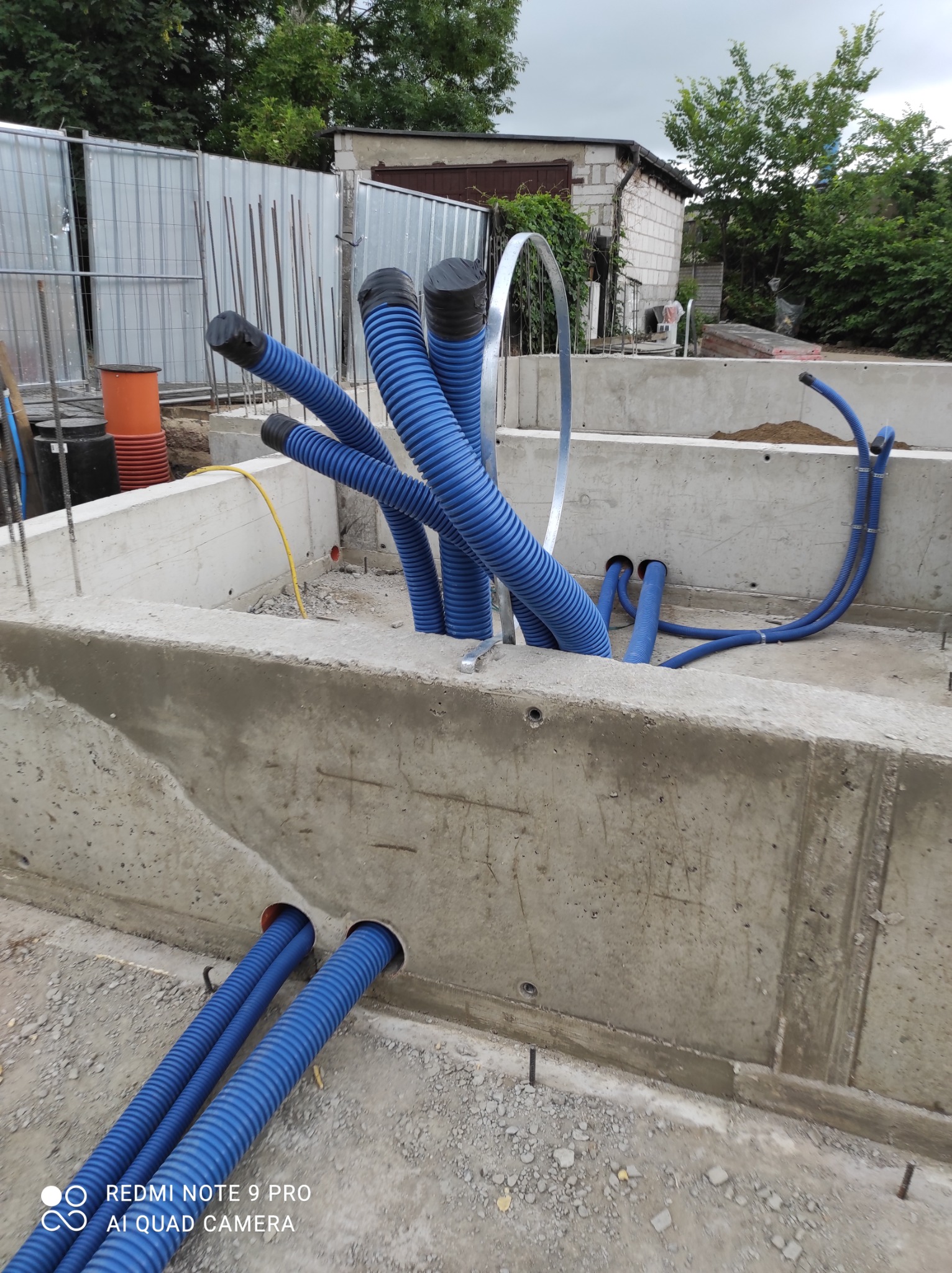 Niebieskie rury karbowane, zabezpieczone czarnymi zaślepkami, wystają z betonowego fundamentu budynku w trakcie budowy. Widoczne elementy szalunku, zbrojenia i inne instalacje.