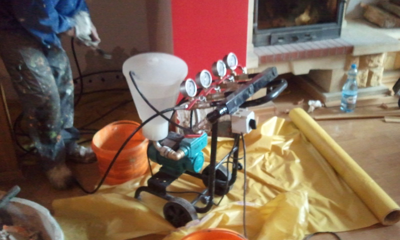 Przenośne urządzenie do iniekcji ciśnieniowej z czterema manometrami, zbiornikiem na płyn i silnikiem elektrycznym, przygotowane do pracy w pomieszczeniu z kominkiem, widoczny pracownik w ubraniu...