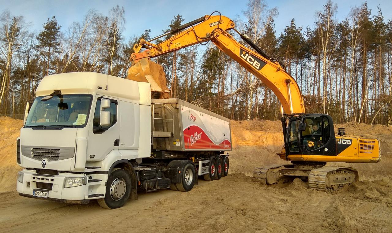 Żółta koparka JCB załadowuje piasek do białej ciężarówki z czerwoną grafiką na naczepie, w tle las.