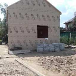 Wznoszony dom szkieletowy z widoczną membraną elewacyjną StroTex 1300, przygotowany do dalszych prac wykończeniowych; na pierwszym planie ułożone płyty chodnikowe i deski.