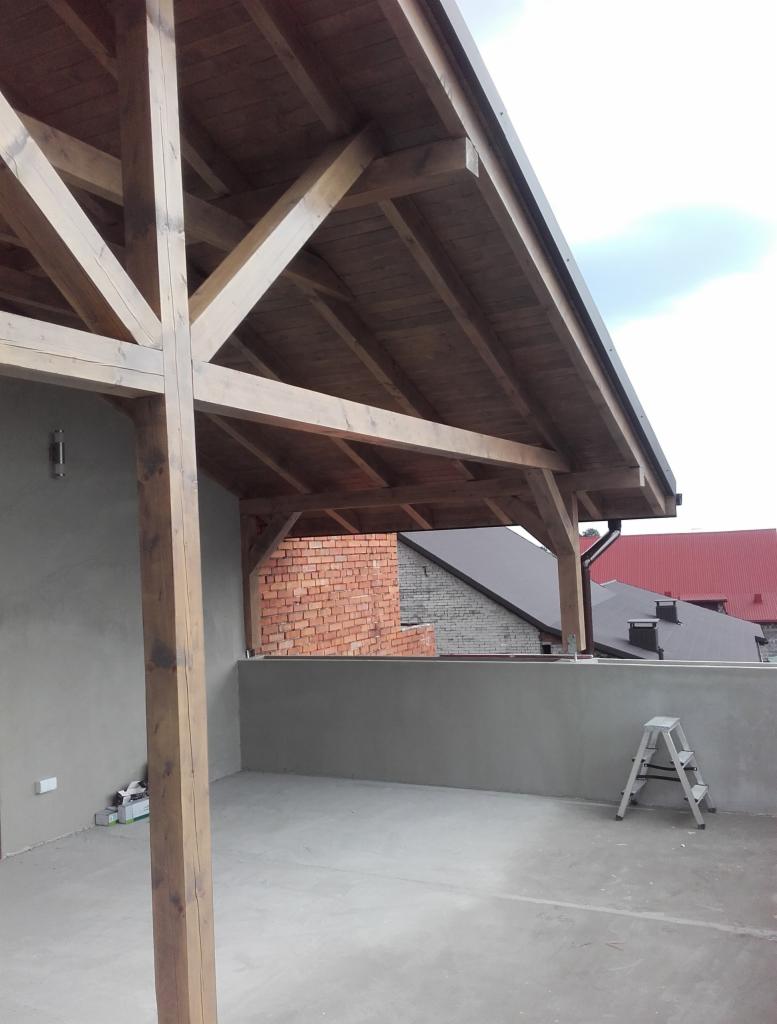 Drewniana konstrukcja zadaszenia poddasza w trakcie budowy, widoczna z zewnątrz, z elementami muru z czerwonej cegły w tle i aluminiową drabinką opartą o ścianę.