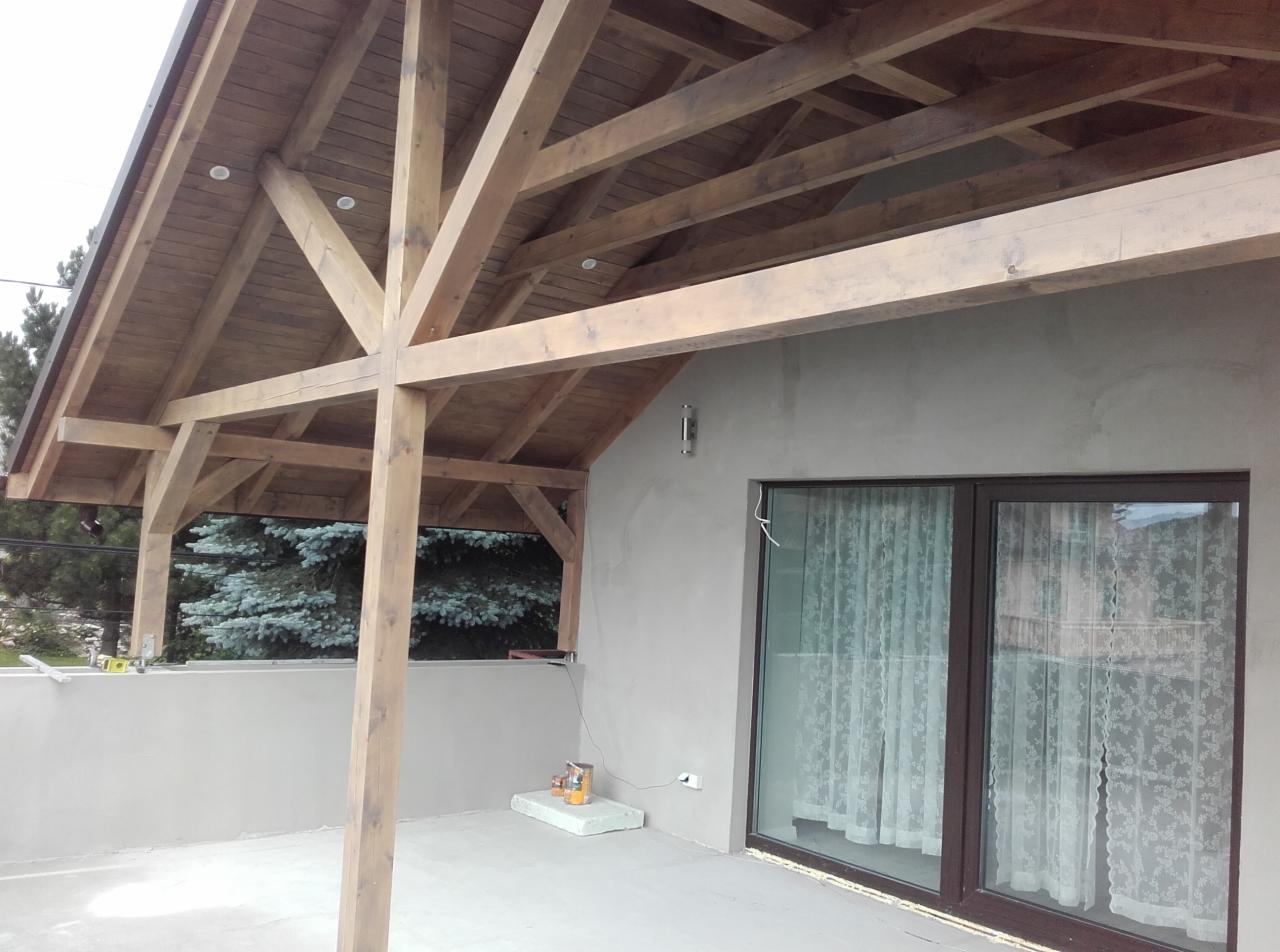 Drewniane zadaszenie poddasza z widoczną konstrukcją szkieletową, oświetleniem punktowym i oknem balkonowym z firankami.