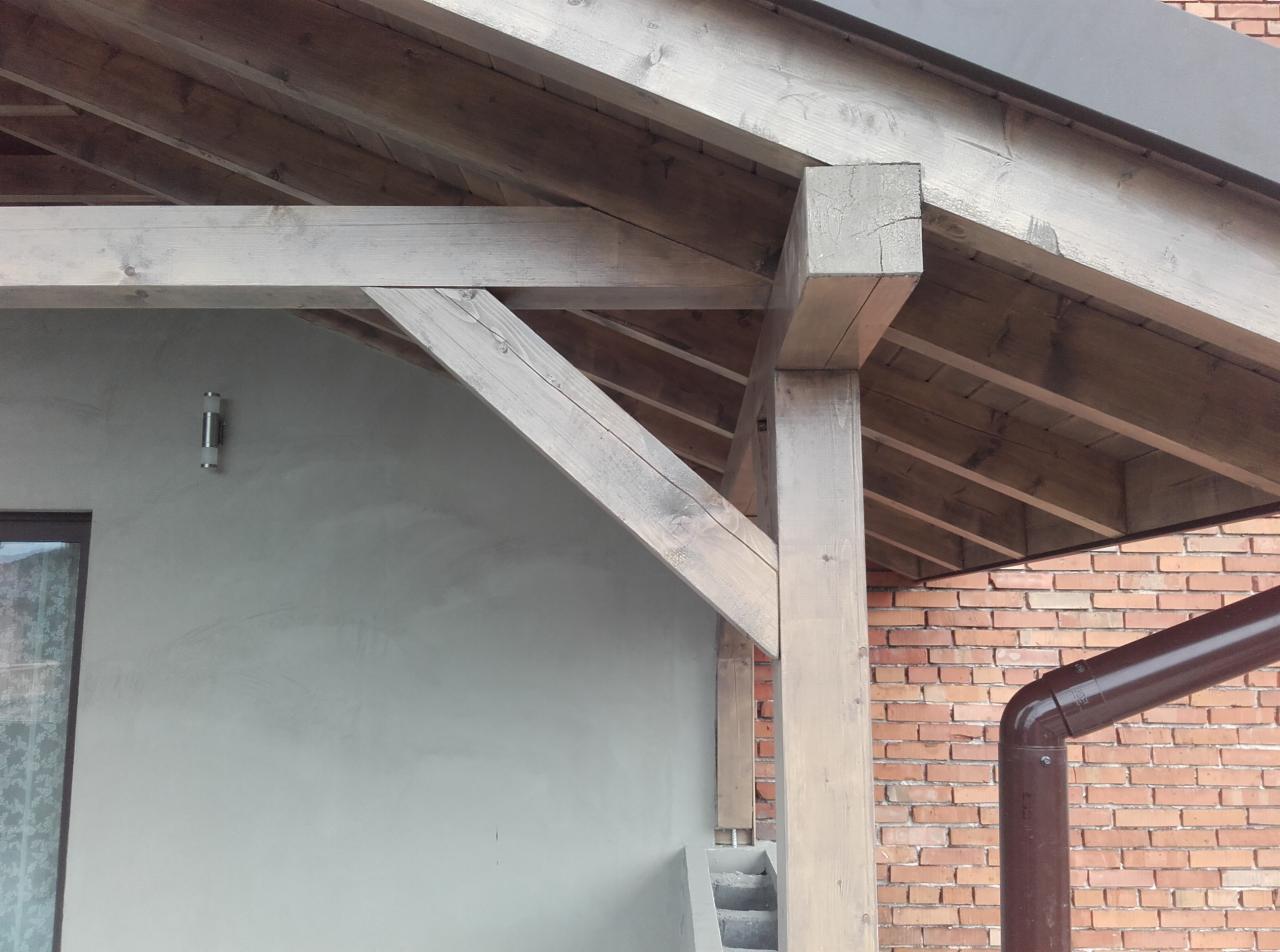 Drewniana konstrukcja zadaszenia poddasza, widoczna na tle otynkowanej ściany i muru z czerwonej cegły, z brązową rynną spustową.