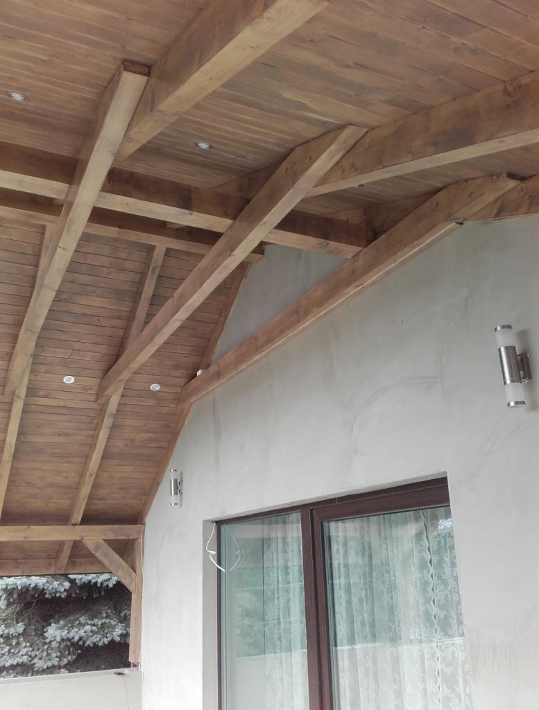 Drewniane zadaszenie tarasu z widocznymi belkami konstrukcyjnymi i wbudowanym oświetleniem punktowym, przylegające do ściany budynku z oknem i lampami zewnętrznymi.