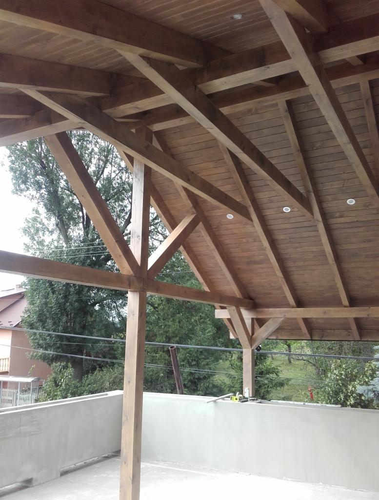 Drewniana konstrukcja zadaszenia poddasza z widocznymi belkami, oświetleniem punktowym i fragmentem otoczenia z zielenią