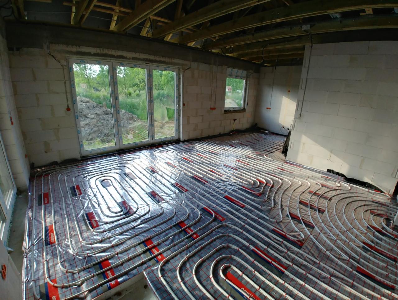 Rozłożone rury ogrzewania podłogowego na folii odblaskowej w surowym wnętrzu domu z oknami wychodzącymi na zieleń.