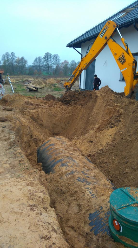 Żółta koparka JCB umieszcza rurę kanalizacyjną w wykopie obok nowego domu z szarym dachem, widoczny zielony właz studzienki.