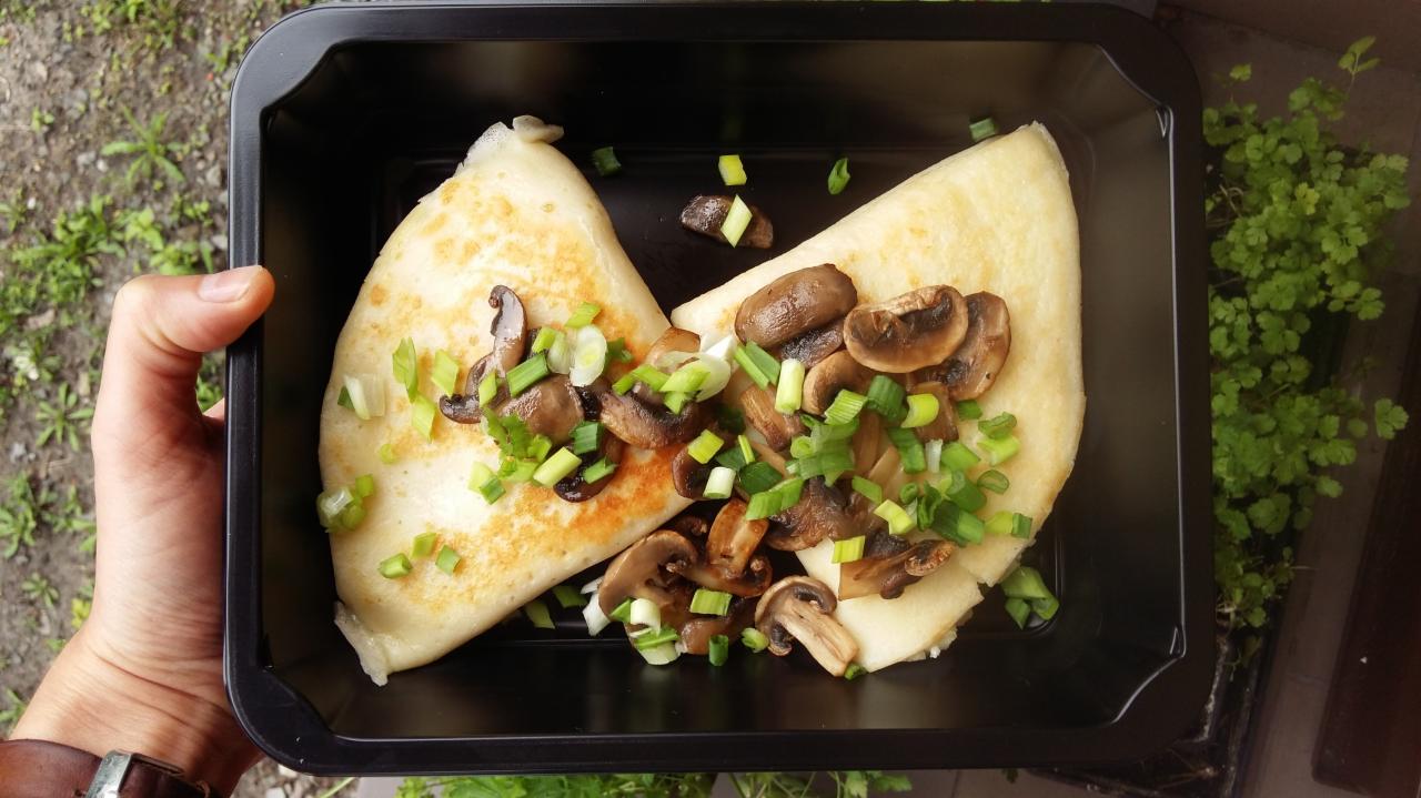 Naleśniki z pieczarkami i szczypiorkiem w czarnym pudełku, trzymane w dłoni, widok z góry.