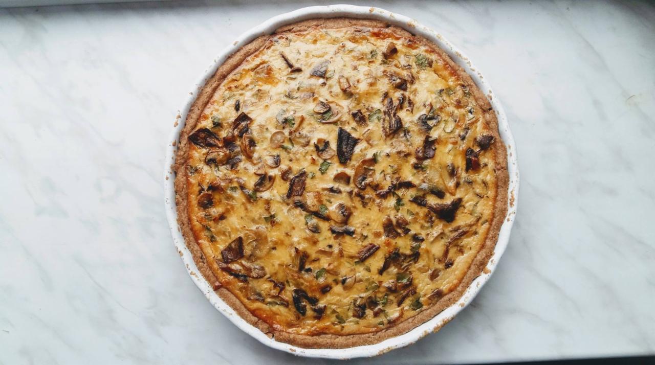 Quiche z pieczarkami i zielonymi ziołami w białej formie na marmurowym blacie, widok z góry.