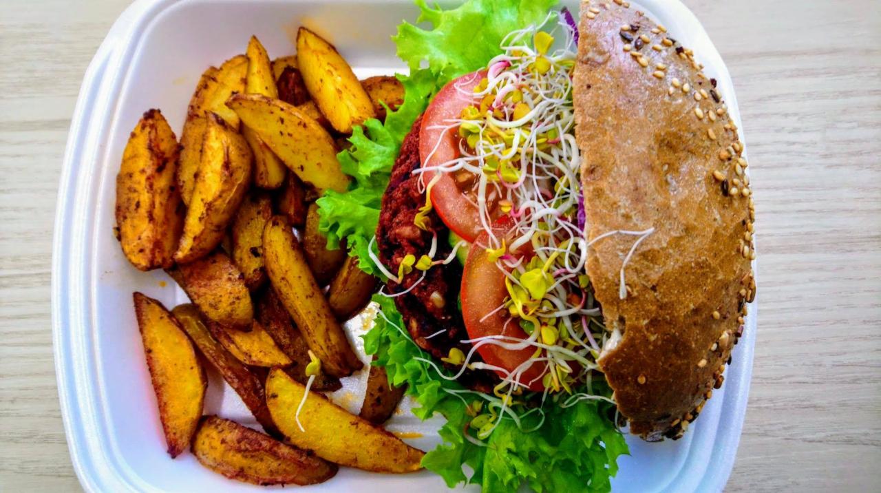 Burger wegetariański z kiełkami i pomidorem obok pieczonych ziemniaków w białym pojemniku.