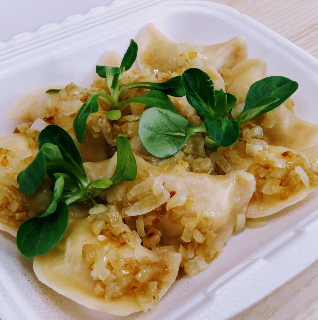 Pierogi z cebulką i świeżą roszponką podane w białym, styropianowym pudełku na wynos.