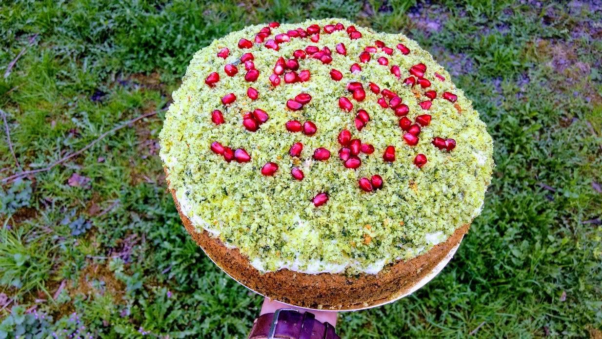Okrągły tort z zieloną posypką i owocami granatu na wierzchu, trzymany na tle trawy.