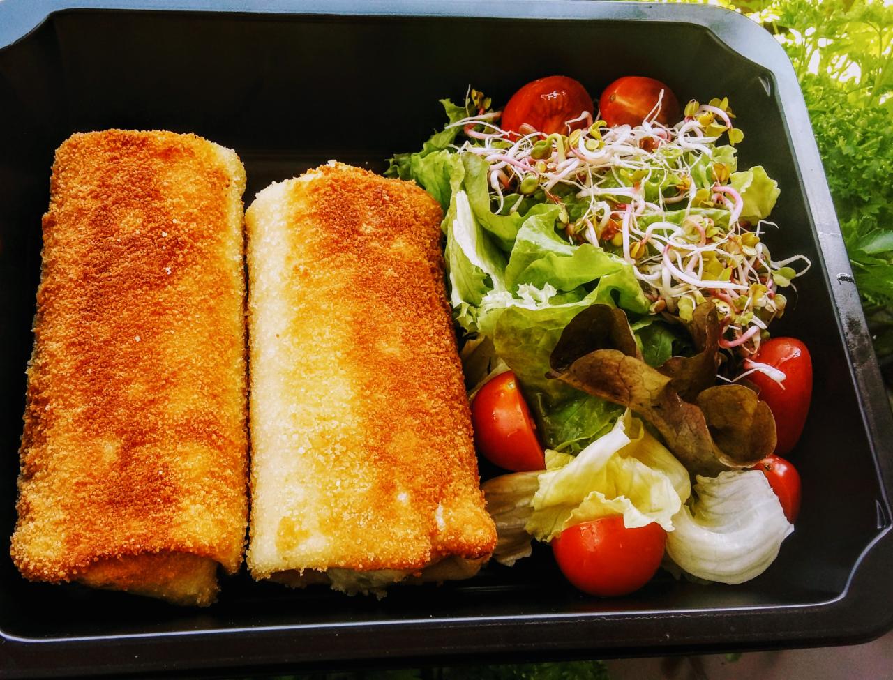 Dwa krokiety z panierką obok sałatki z pomidorami cherry i kiełkami rzodkiewki w czarnym pojemniku cateringowym.