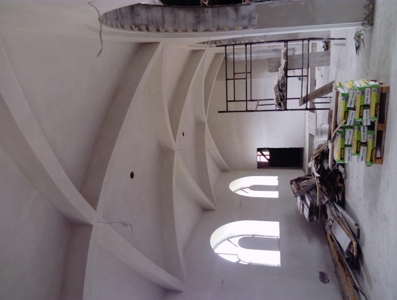 Wnętrze budynku w trakcie prac: surowy tynk na sklepieniu krzyżowym, widoczne okna łukowe, rusztowanie, paleta z workami zaprawy i porozrzucane deski.