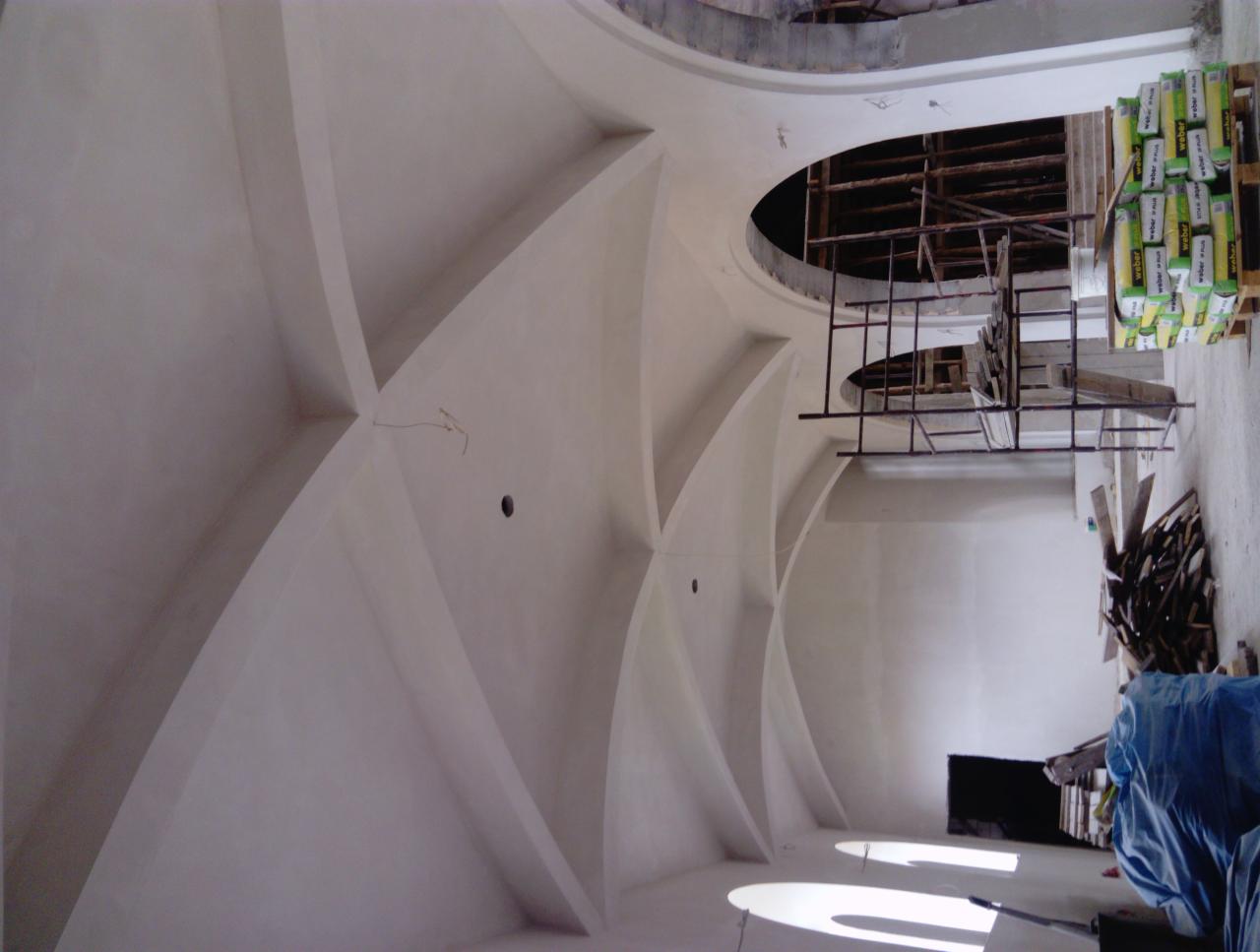 Wnętrze w trakcie prac wykończeniowych: świeżo otynkowany sufit z dekoracyjnymi łukami, widoczne rusztowania i paleta z workami zaprawy.