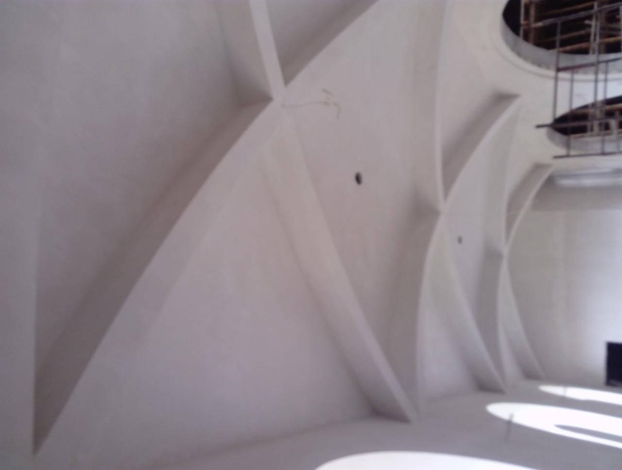 Wnętrze z surowymi, białymi ścianami i dekoracyjnymi, łukowatymi elementami architektonicznymi, widoczne otwory w suficie i fragment rusztowania.