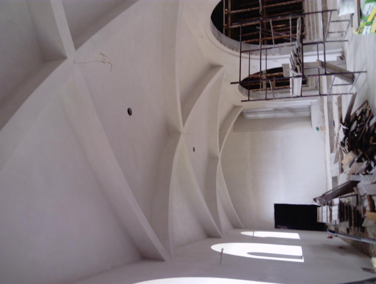 Wnętrze w trakcie wykańczania: sufit z dekoracyjnymi łukami, widoczne rusztowania i materiały budowlane, światło wpadające przez okna.