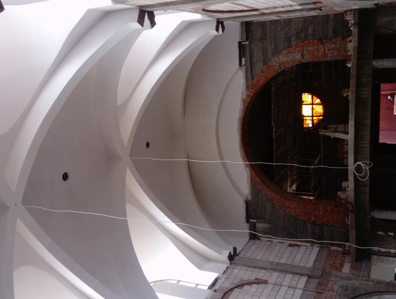 Wnętrze budynku w trakcie remontu, widoczne sklepienie łukowe po tynkowaniu, fragment muru z cegły wokół okna z witrażem oraz elementy konstrukcyjne i rusztowania.