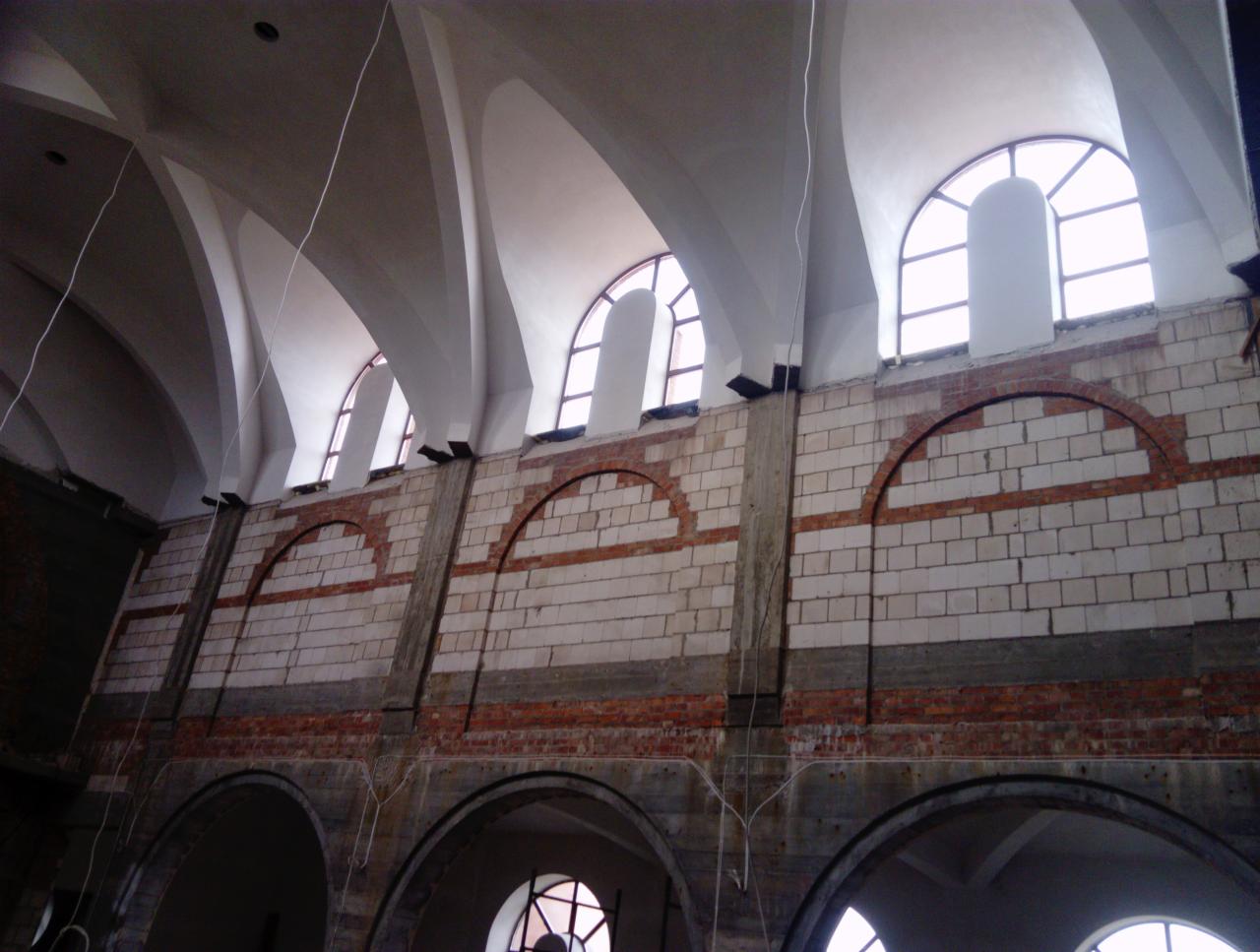 Wnętrze budynku w trakcie renowacji, widoczne ceglane ściany z łukowymi oknami i sklepieniem, częściowo odnowione, z widocznymi śladami wcześniejszych prac.