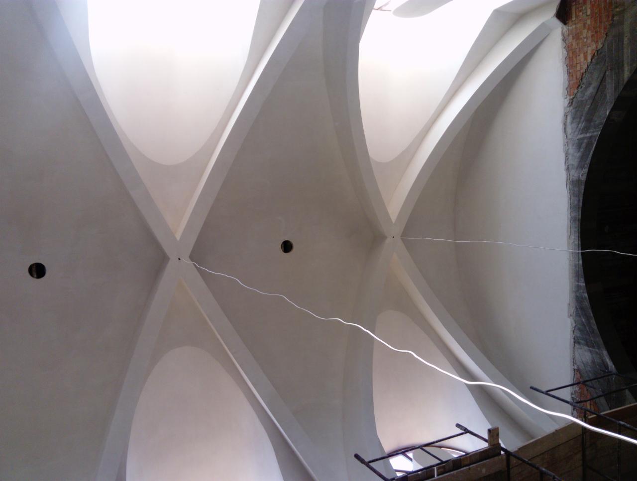 Krzyżowe sklepienie łukowe z widocznymi otworami na oświetlenie, świeżo otynkowane na gładko, z fragmentem muru z cegły i rusztowaniem w tle.