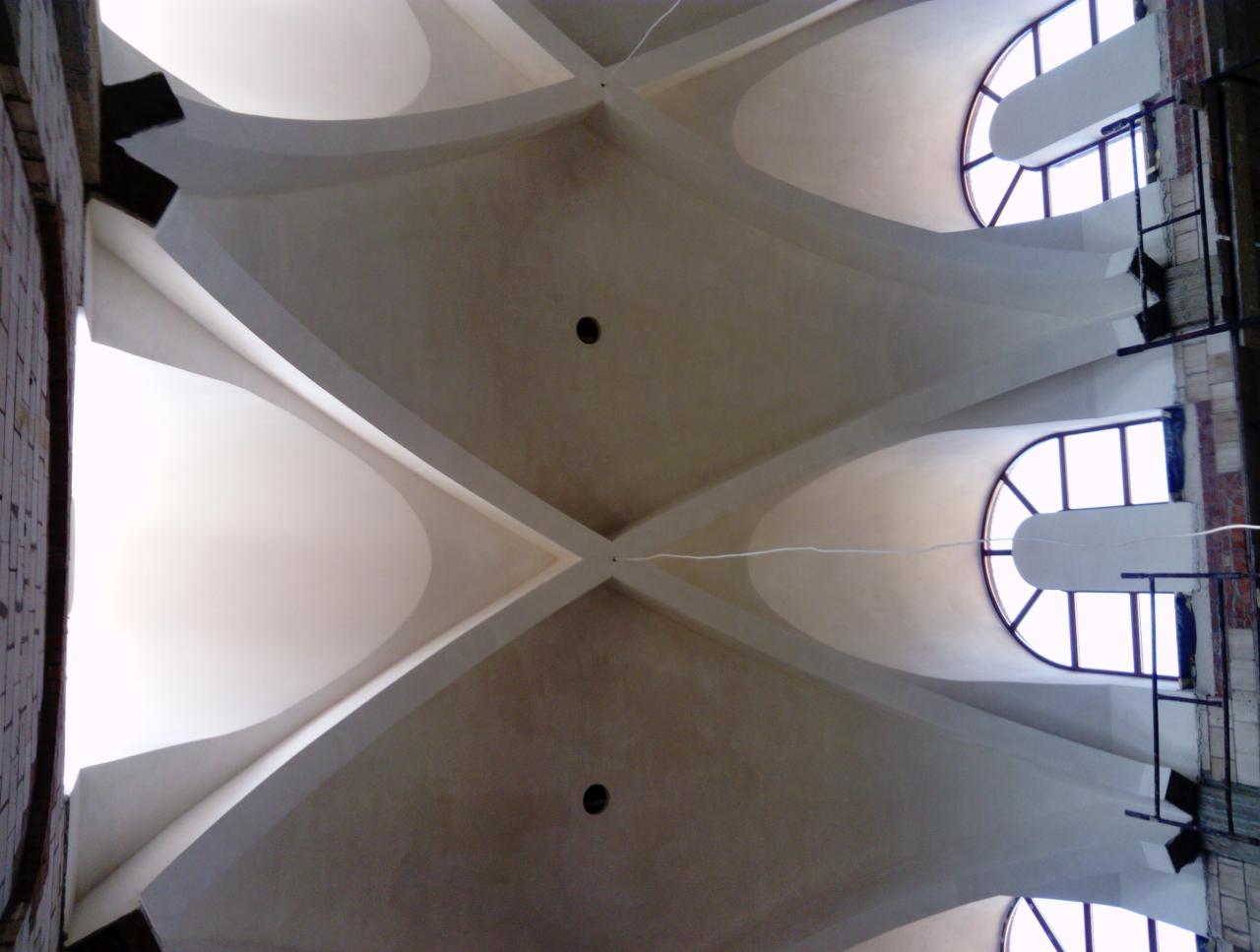 Skomplikowana struktura sufitu z krzyżującymi się łukami, widoczne okna łukowe, surowe wykończenie ściany z cegły i fragment rusztowania.