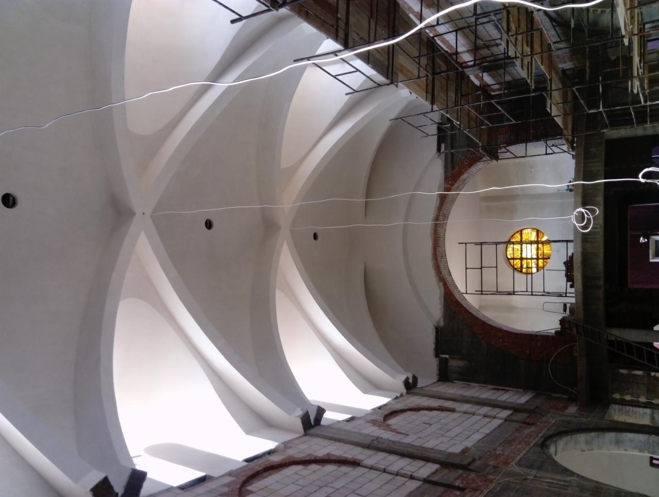 Wnętrze zabytkowego budynku w trakcie prac renowacyjnych, widoczne sklepienie krzyżowe z nowym tynkiem, okrągłe okno z witrażem i rusztowania.