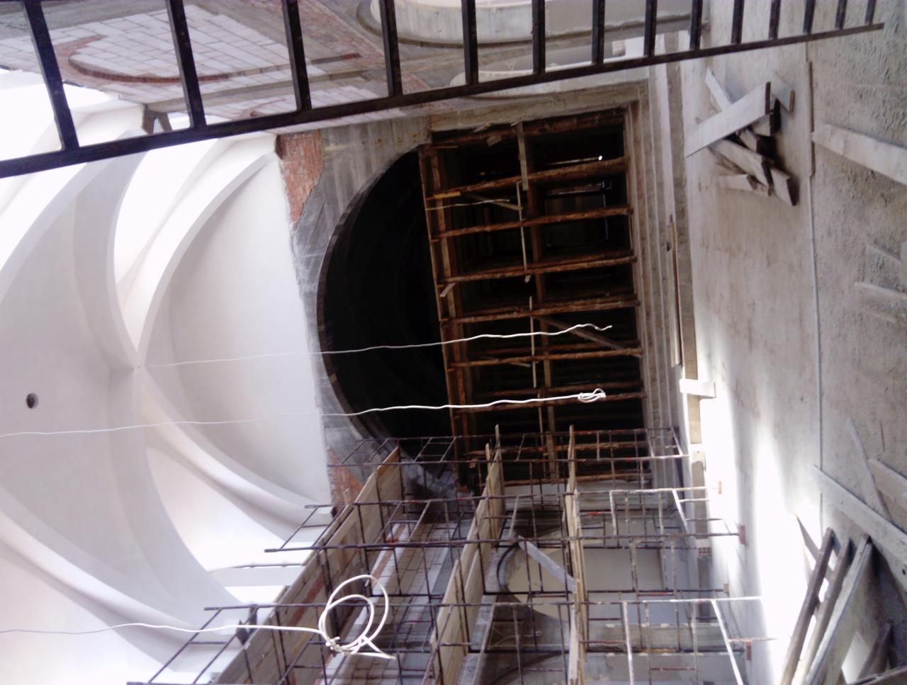 Wnętrze w trakcie budowy z widocznym rusztowaniem, fragmentem sklepienia z dekoracyjnymi łukami i otworem w ścianie z ceglanym obramowaniem.