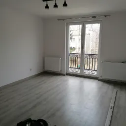 Wykończony pok&oacute;j z szarymi panelami podłogowymi, białymi ścianami i grzejnikami, czarną lampą sufitową i balkonem z ozdobną balustradą. Na podłodze widoczny odkurzacz i listwa przypodłogowa.