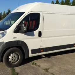 Biały samochód dostawczy Fiat Ducato na betonowym podłożu, z widocznymi czerwonymi siedzeniami przez szybę i zielonymi drzewami w tle.