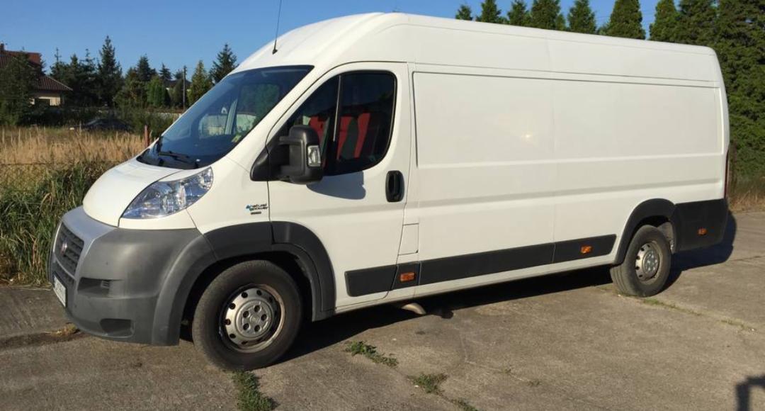 Biały samochód dostawczy Fiat Ducato na betonowym podłożu, z widocznymi czerwonymi siedzeniami przez szybę i zielonymi drzewami w tle.