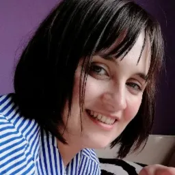 Portret uśmiechniętej kobiety w bluzce w niebiesko-białe paski na tle fioletowej ściany, z widocznym fragmentem czarno-białej poduszki.