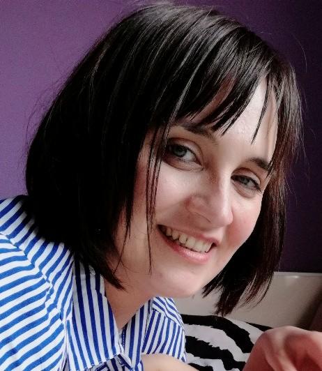 Portret uśmiechniętej kobiety w bluzce w niebiesko-białe paski na tle fioletowej ściany, z widocznym fragmentem czarno-białej poduszki.