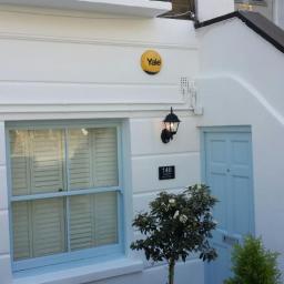 Propainting Sp z.o.o - Biała fasada domu z błękitnymi akcentami: okno z żaluzjami, drzwi wejściowe, lampa ścienna i tabliczka z numerem 146, uzupełniona małym drzewkiem i logo Yale.