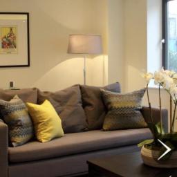 Propainting Sp z.o.o - Jasne wnętrze salonu po remoncie: szary narożnik z wzorzystymi poduszkami i żółtą poduszką, lampa podłogowa, storczyk w doniczce na stoliku kawowym, oświetlenie sufitowe.