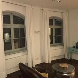 Propainting Sp z.o.o - Wnętrze pokoju po remoncie: dwa okna z szarymi ramami i białymi zasłonami, brązowa skórzana sofa, stolik kawowy i turkusowa konsola z dekoracyjnymi wazonami.