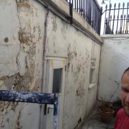 Propainting Sp z.o.o - Zniszczona elewacja zewnętrzna budynku z widocznymi odpadającymi fragmentami farby i tynku, przygotowana do renowacji, z fragmentem rusztowania w kadrze i widoczną częścią osoby w czerwonej koszulce.