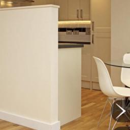 Propainting Sp z.o.o - Jasne, kremowe ściany w odnowionej kuchni z drewnianą podłogą i nowoczesnymi białymi krzesłami przy szklanym stole, widok na zabudowę kuchenną z oświetleniem podszafkowym.