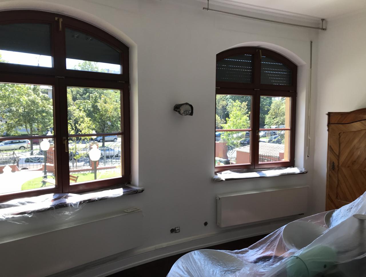 Pokój z dwoma oknami łukowymi zabezpieczonymi folią malarską, ściany świeżo pomalowane na biało, widoczny fragment mebla pod folią ochronną.