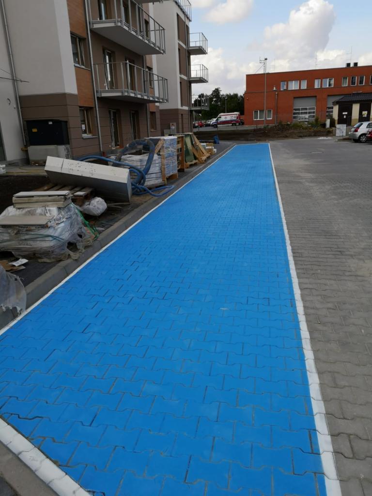 Świeżo ułożona, jaskrawo niebieska kostka brukowa na chodniku przed nowym budynkiem mieszkalnym, z widocznymi materiałami budowlanymi w tle.