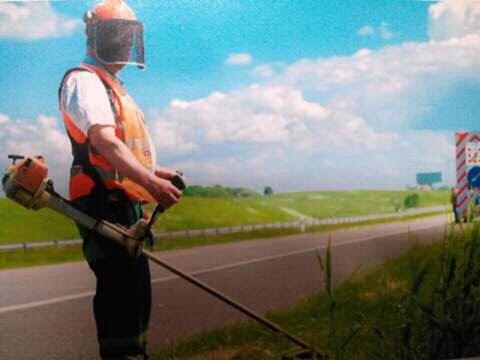 Pracownik w kasku i kamizelce odblaskowej, z podkaszarką spalinową przy drodze, koszenie trawy na poboczu, w tle zielone pola i błękitne niebo z chmurami.