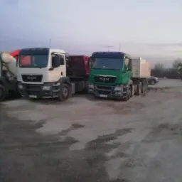 Dwa samochody ciężarowe MAN: biały betonomieszarka i zielony wywrotka, stojące na placu budowy o zmierzchu. Widoczne ślady opon na ziemi.