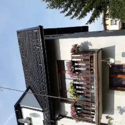 Dom z nowym dachem pokrytym czarną blachodachówką, widoczne obróbki blacharskie, antena satelitarna i balkony z kwiatami.