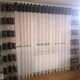 Zasłony w oknie: półprzezroczysta siatka z brązowymi, dekoracyjnymi pasami u góry i dołu, zawieszone na ciemnobrązowych kółkach