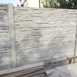 Szary betonowy płot z fakturą kamienia, widoczne elementy montażowe i fragmenty drewnianych desek u podstawy.