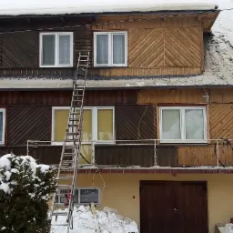 Dwukondygnacyjny dom z drewnianą elewacją w trakcie czyszczenia, widoczna drabina oparta o ścianę, część elewacji po lewej stronie ciemniejsza, po prawej jaśniejsza, zimowy krajobraz z dużą ilością...