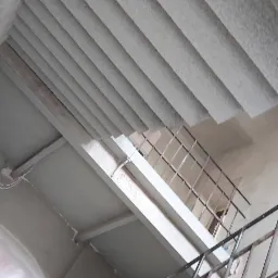 Klatka schodowa w trakcie remontu, widoczne surowe betonowe schody i metalowa balustrada, ślady po skuwanym tynku na ścianach.