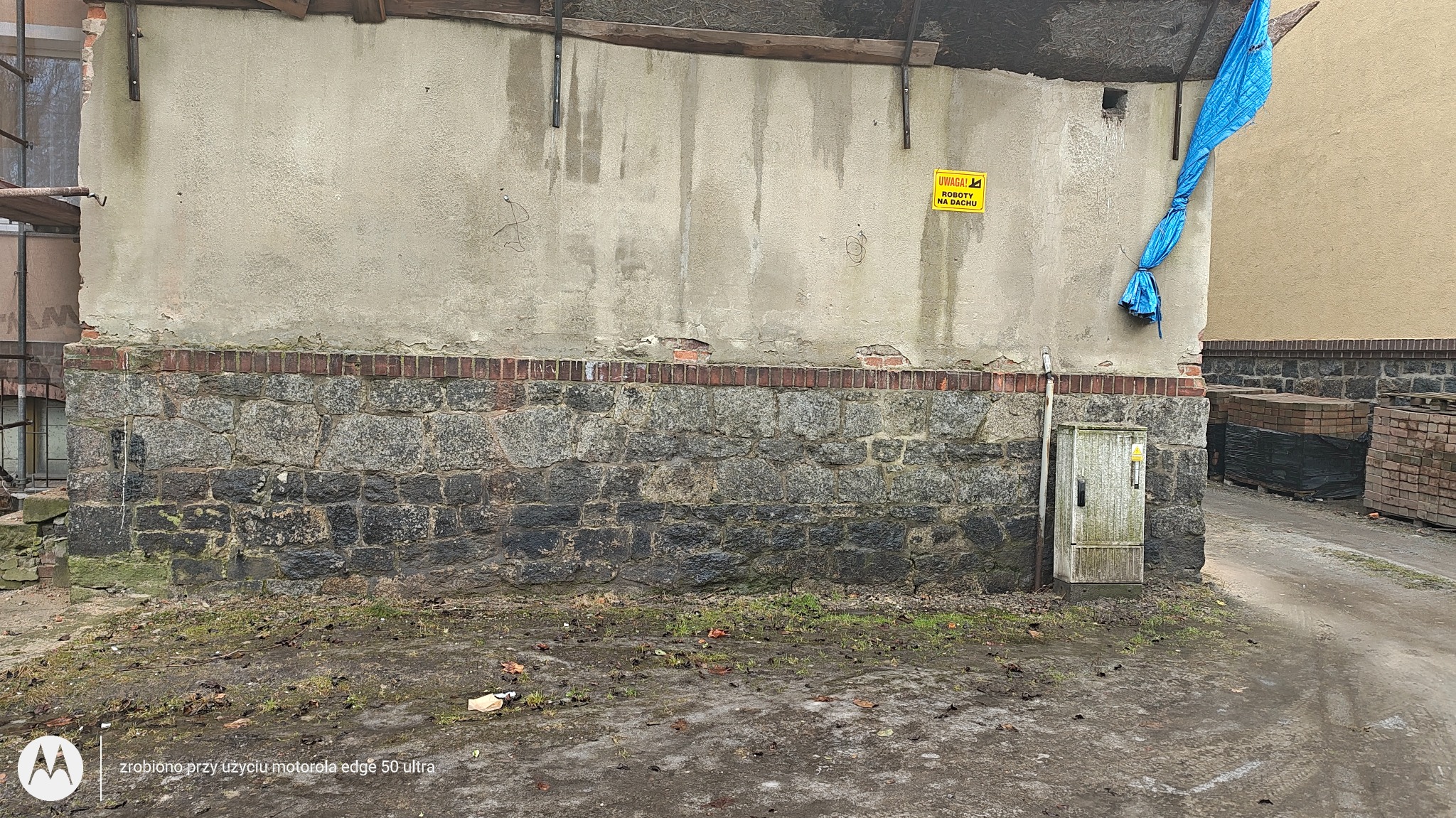 Murek z kamienia i cegły z ubytkami, fragment ściany z żółtą tabliczką ostrzegawczą i niebieską plandeką. Widoczne ślady wilgoci i zabrudzeń.
