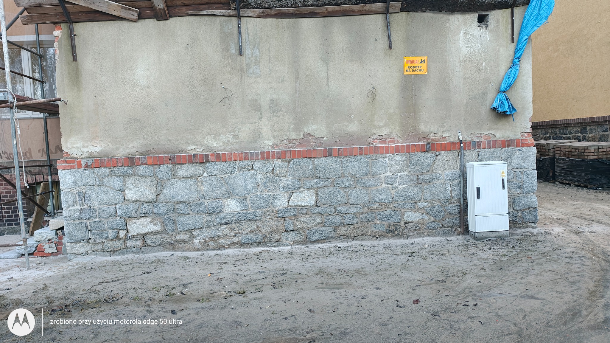 Fragment elewacji budynku z kamienną podmurówką i fragmentem rusztowania. Widoczny także fragment instalacji elektrycznej i tabliczka ostrzegawcza.