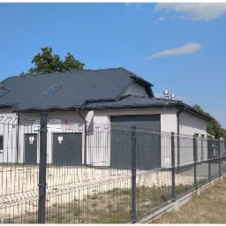 Nowoczesny budynek gospodarczy z szarą blachą trapezową na dachu, widoczny zza metalowego ogrodzenia, z zielonym zbiornikiem w tle.