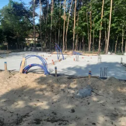 Przygotowany fundament pod budowę domu jednorodzinnego, widoczne wystające pręty zbrojeniowe oraz instalacja wodna w niebieskich rurach, tło stanowią wysokie sosny.